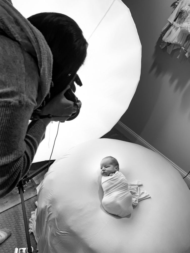Overhead view of photographer Yvonne Leon photographing a wrapped newborn on a posing beanbag; studio light to her left.