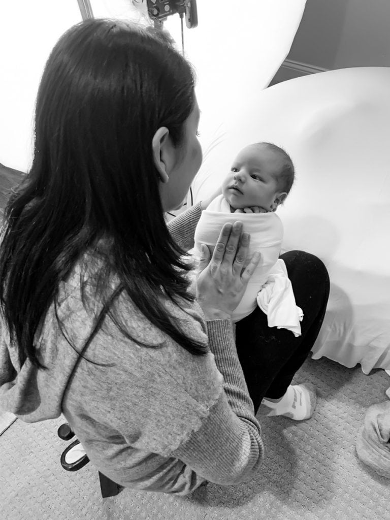 Photographer Yvonne Leon in studio holding a wrapped newborn, her back to the camera; relaxed baby gazes at her.