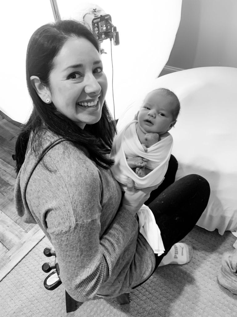 Photographer Yvonne Leon sits in studio, smiling and showing a swaddled newborn toward the camera; studio light behind them.