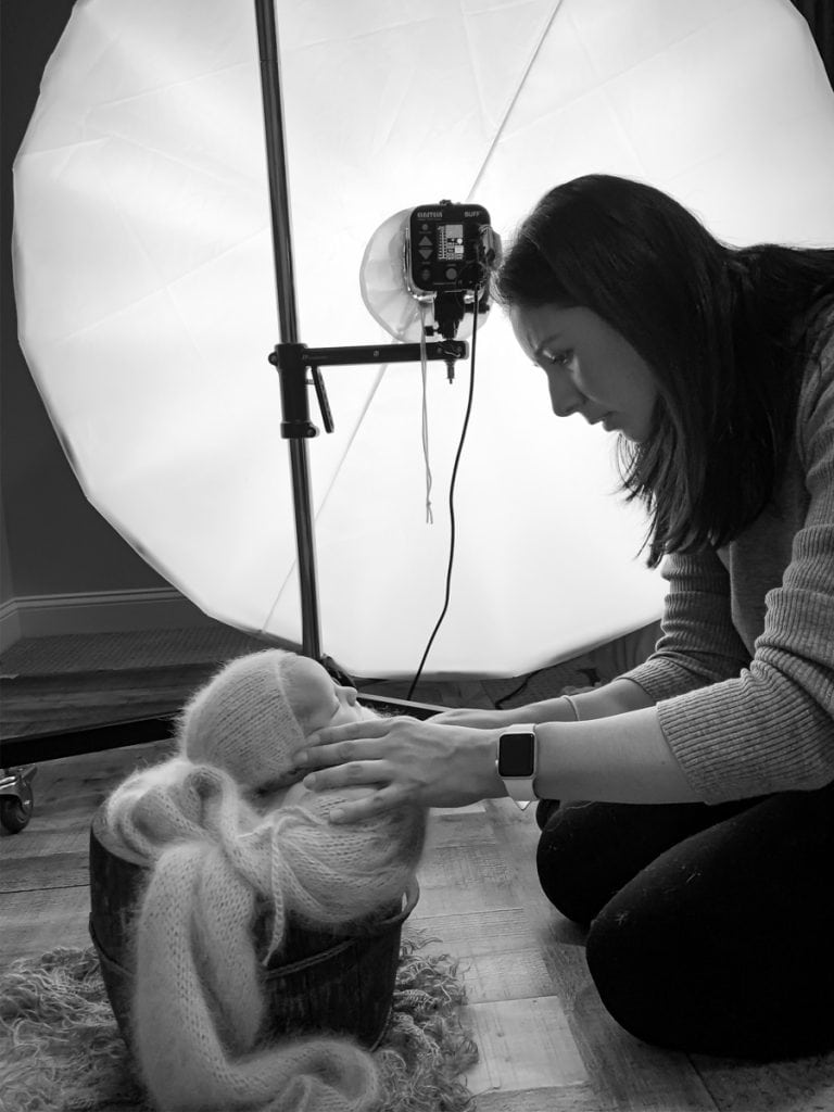 Photographer Yvonne Leon posing newborn in bucket prop on the floor during a studio session; studio light in background.