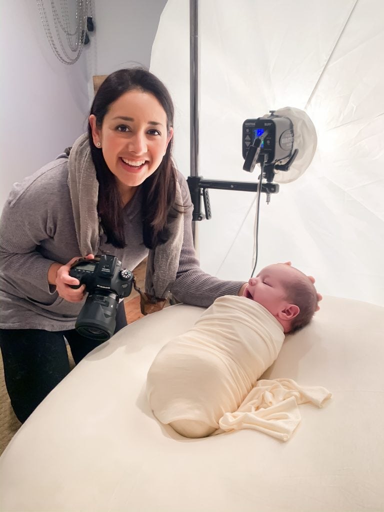 Photographer Yvonne Leon smiling while holding camera; standing next to wrapped newborn laying on a beanbag in studio.