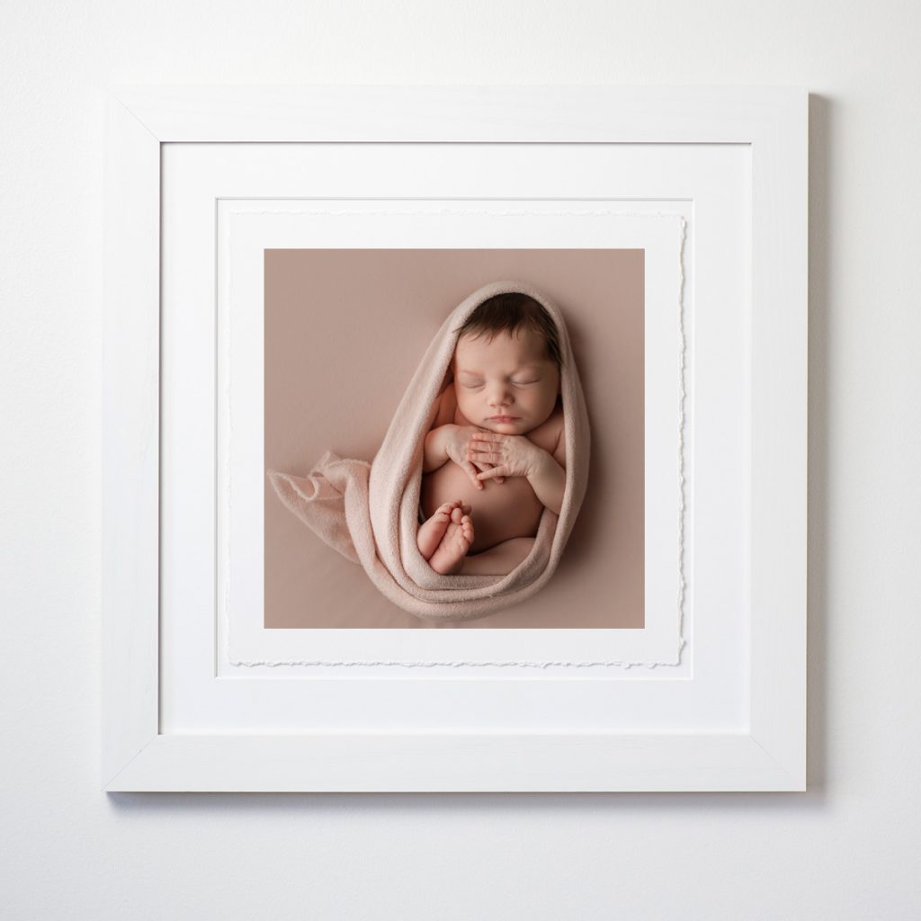 White square frame with deckled-edge fine art print of newborn sleeping on pink backdrop and wrap, showcasing studio artwork.