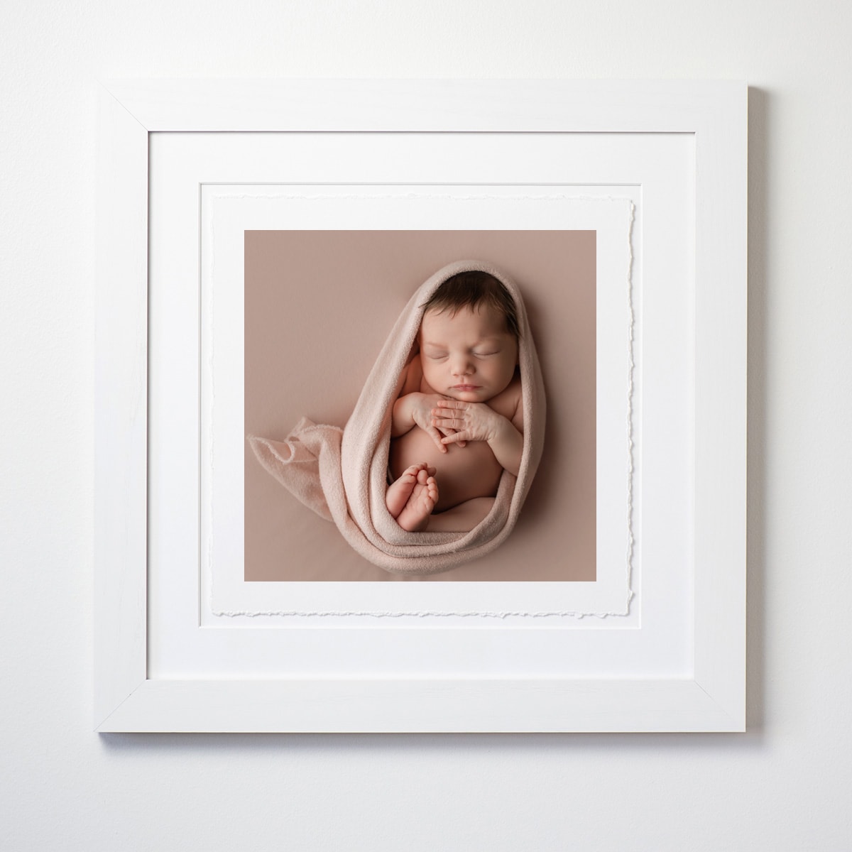 White square frame with deckled-edge fine art print of newborn sleeping on pink backdrop and wrap, showcasing studio artwork.