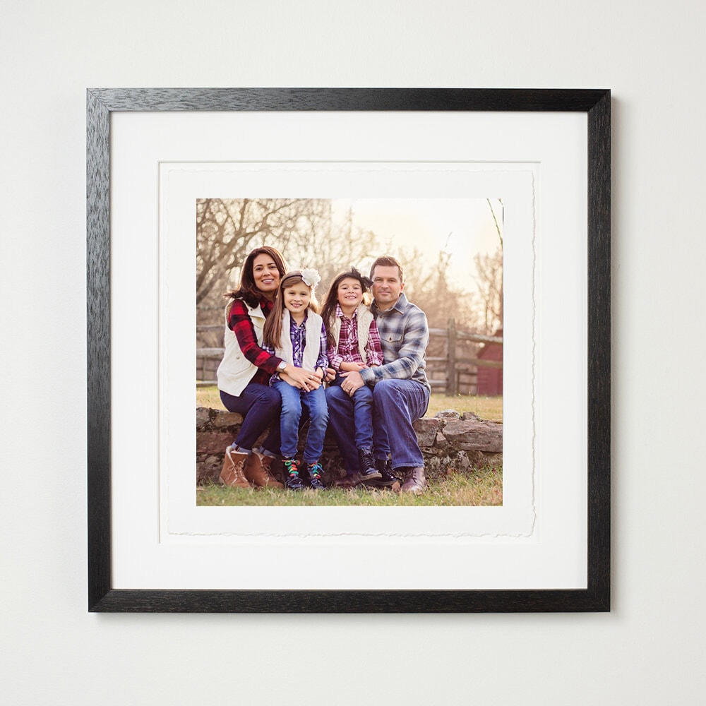 Framed matted portrait of mom, dad, and two daughters sitting close in outdoor fall photo; showcasing studio artwork.