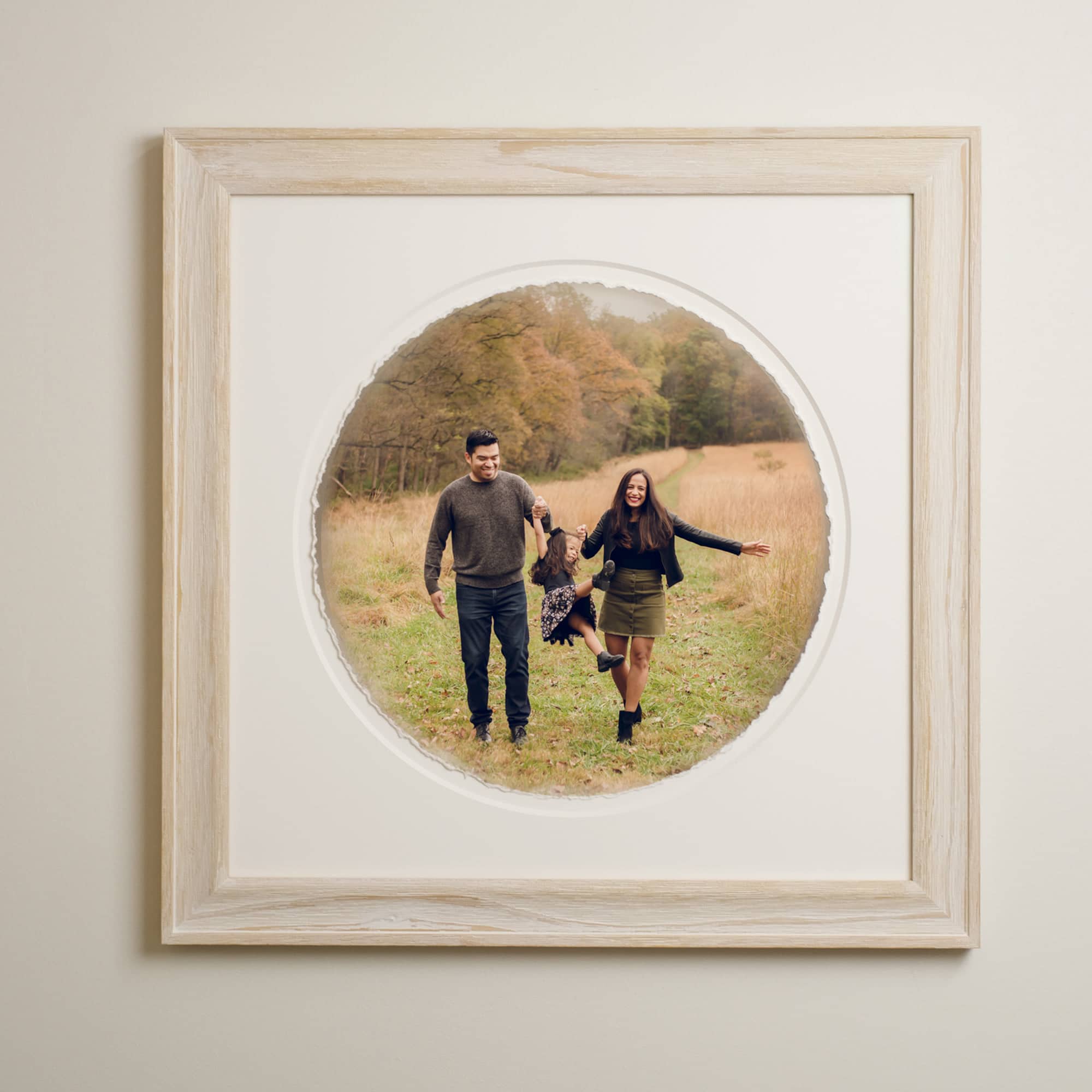 Circle-shaped portrait of parents swinging toddler daughter in grassy field; matted and displayed in a white-wash oak frame.