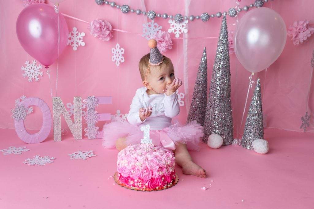 baby girl with a pink tutu skirt on eating pink cake with snowflake sprinkles on top for a wintery birthday theme.