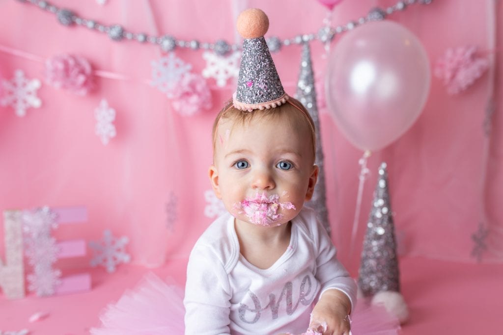 closeup of baby girl's face covered in pink frosting from eating birthday cake during a first birthday cake smash photoshoot.