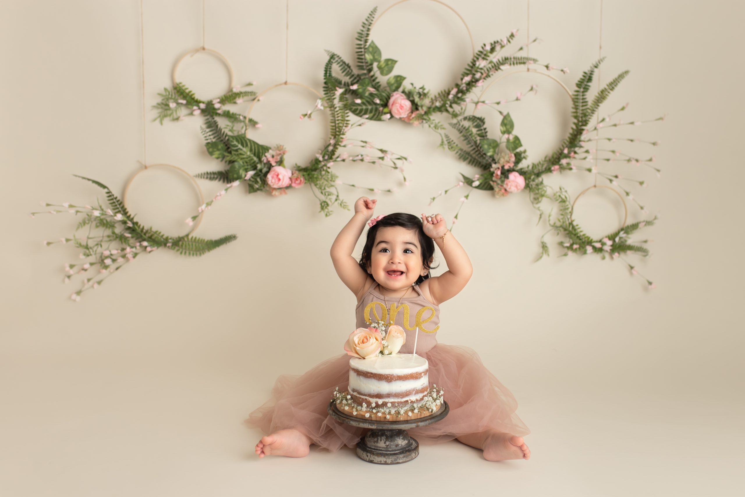 Happy baby girl in pink dress with arms raised and big smile; floral hoops and beige backdrop in studio cake smash session.