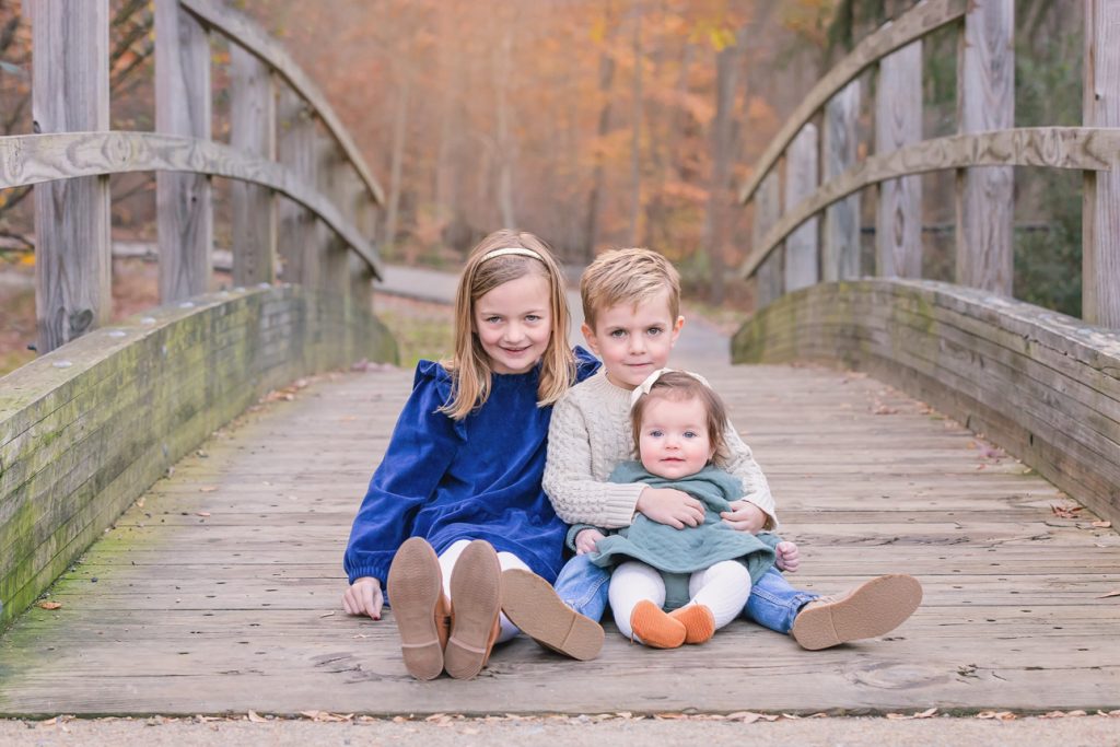 Three young siblings sit on wooden bridge during a fall family session; boy holds infant sister; colorful foliage background.