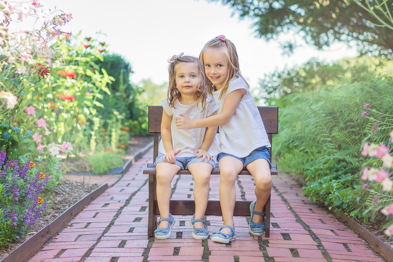 Two young sisters in matching outfits sit on small bench on a brick walkway surrounded by spring flower beds, outdoor session.