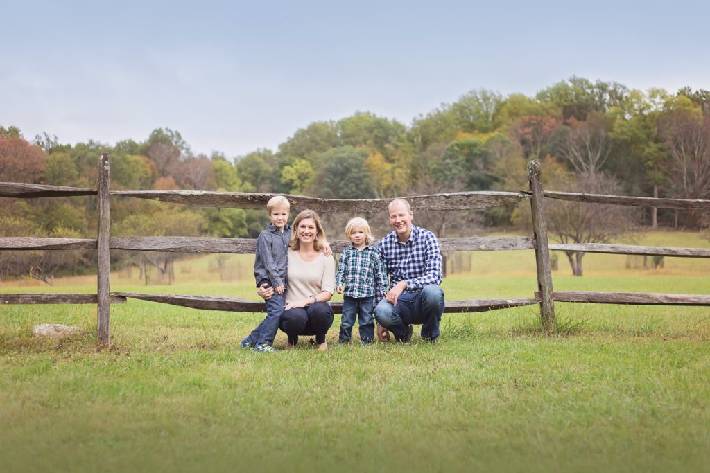 Family of four with two young sons stand on grass by wooden fence, trees in distance with early foliage; all smiling.