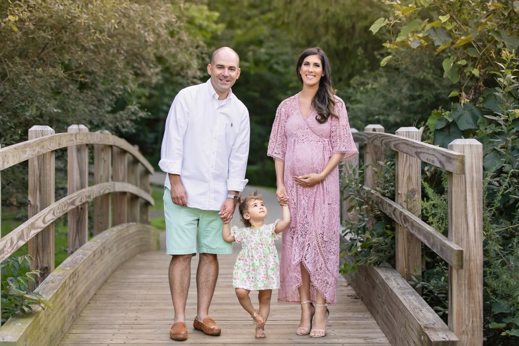 Family of three standing on wooden bridge; mom and dad holding toddler girl's hands as she looks up at mom's pregnant belly.