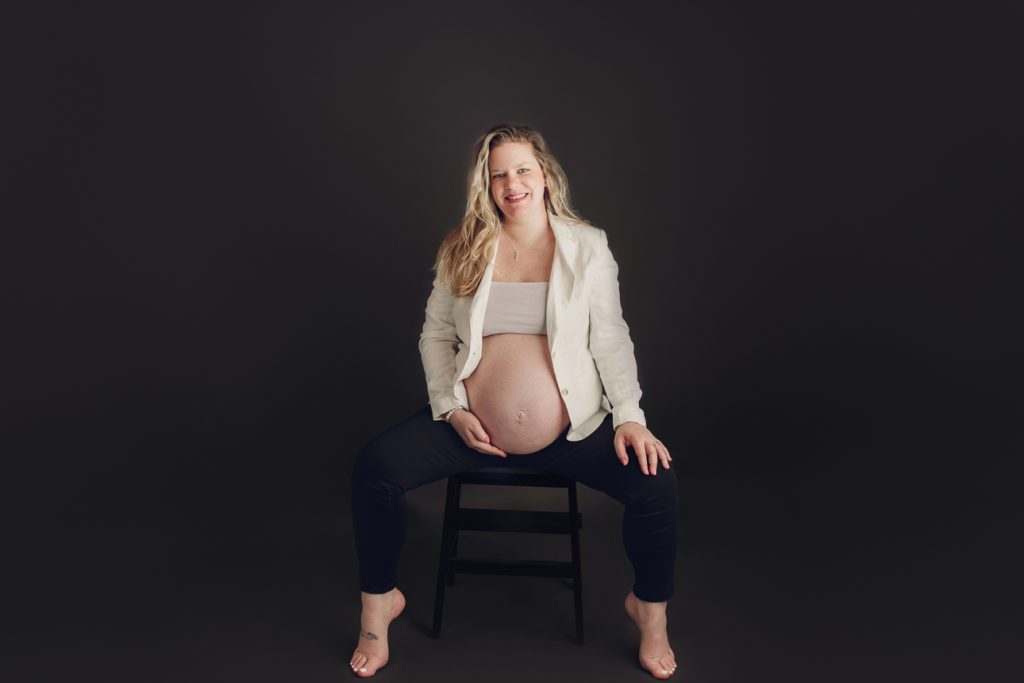 Maternity studio portrait of mom sitting on stool wearing open jacket exposing belly; black background for dramatic effect.