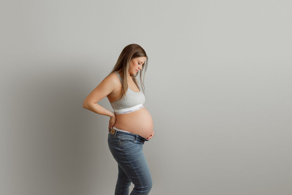 Studio maternity portrait on grey backdrop, mom wearing Calvin Klein bra and unbuttoned jeans with exposed belly.