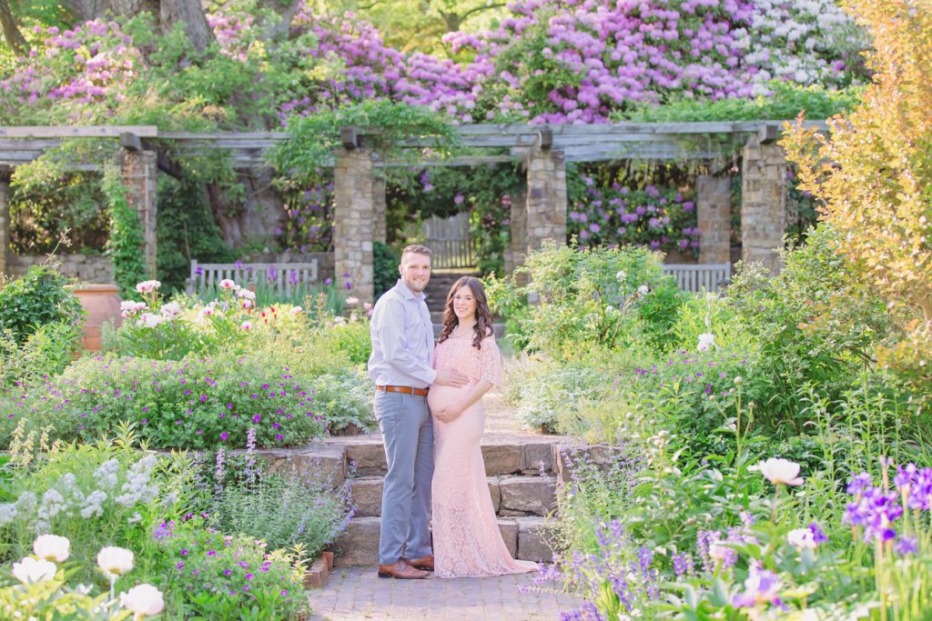 Maternity couple in elegant attire standing in spring garden with pergola surrounded by blooms; smiling and holding belly.