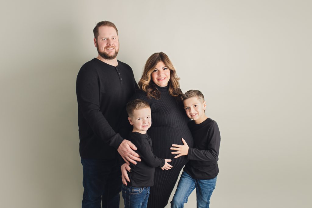 Studio maternity portrait of expecting mom, dad and two young boys all dressed in black; holding each other and mom's belly.