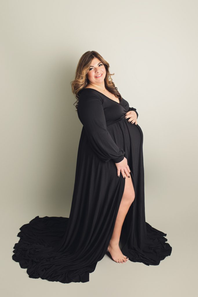 Studio maternity portrait in long black dress with one hand on belly; styled hair and makeup; light grey backdrop.