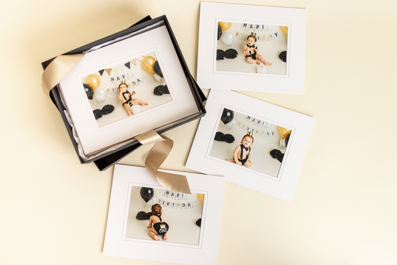 Flat lay of baby boy cake smash matted portraits displayed in a black gift box, showcasing professional print products.