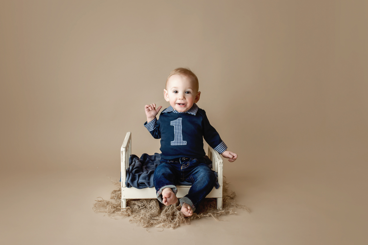 Smiling baby boy sitting in a little bed prop wearing a blue onesie with number 1 during a first birthday milestone session.
