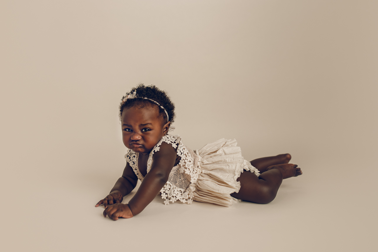 Baby girl with dark skin in cream romper lying on tummy, making a cute pouty face at the camera during milestone session.