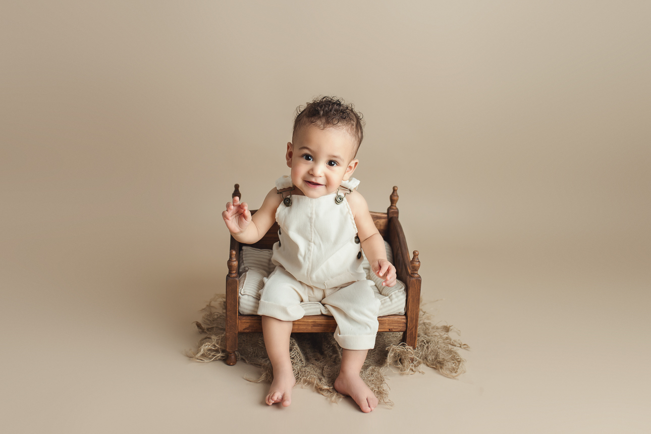 Smiling baby boy in cream-colored overalls sitting in wooden bed prop during a studio milestone photo session.