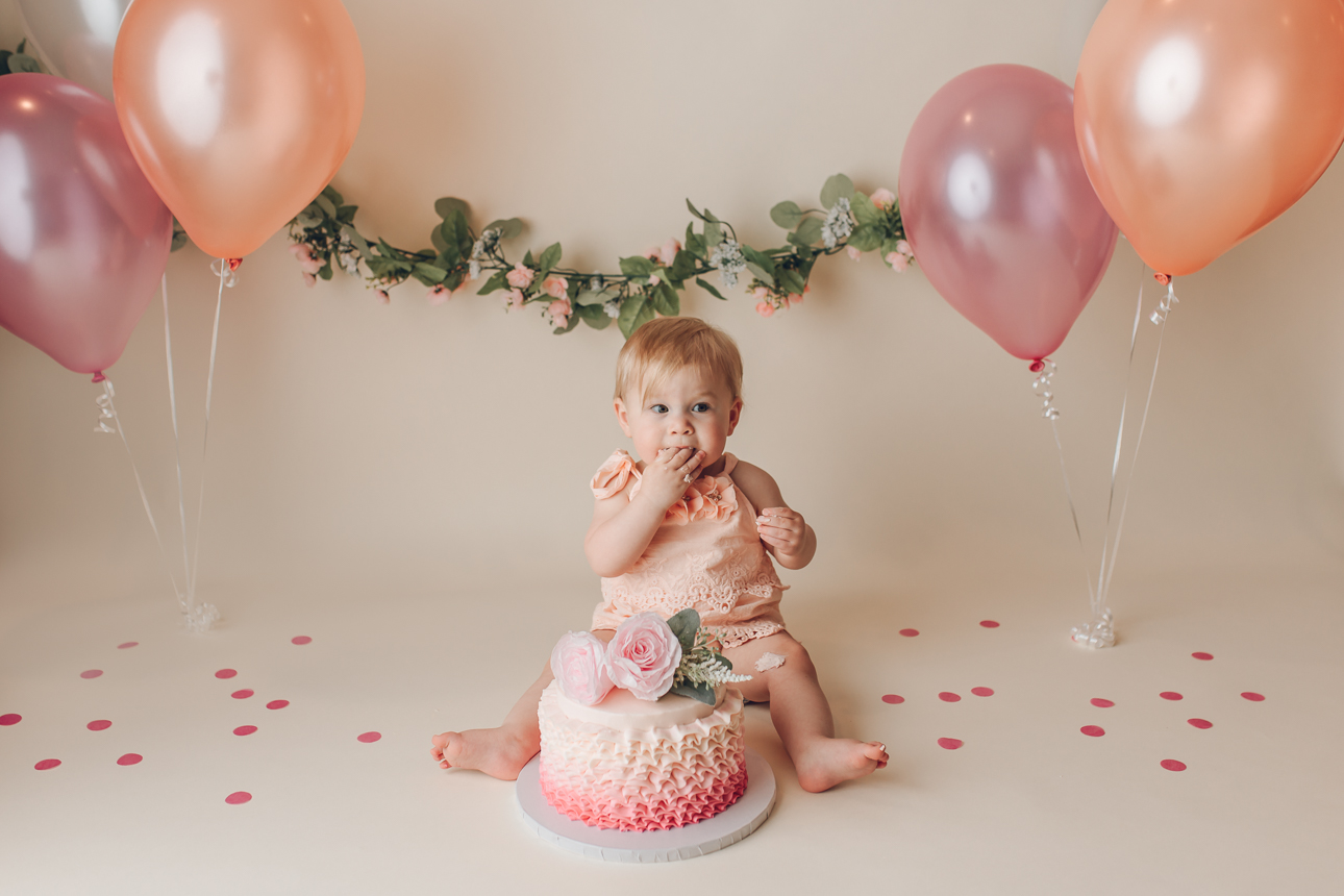 Baby girl in peach romper sitting and eating cake during cake smash session; floral garland and balloons on cream backdrop.