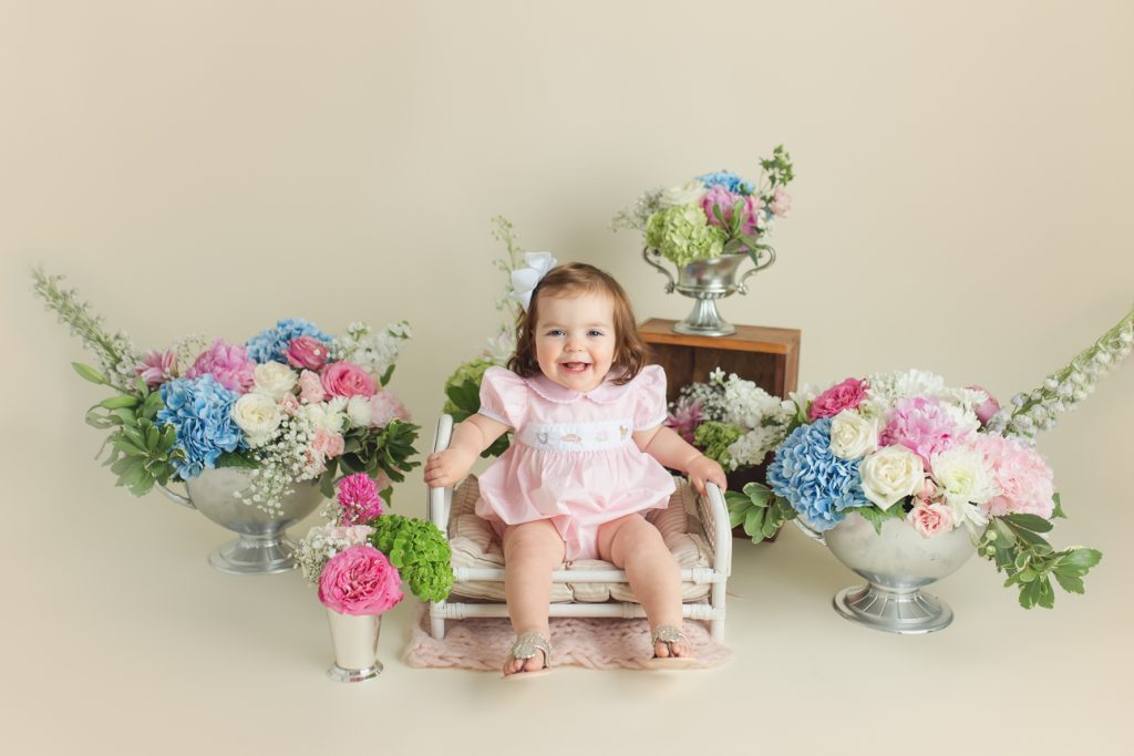 Smiling baby girl in pink dress surrounded by pink, blue, and green flowers during floral-themed cake smash photo session.