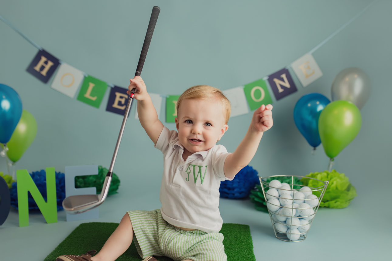 Smiling baby boy in a golf-themed cake smash session, sitting up and holding a mini golf club while looking at the camera.