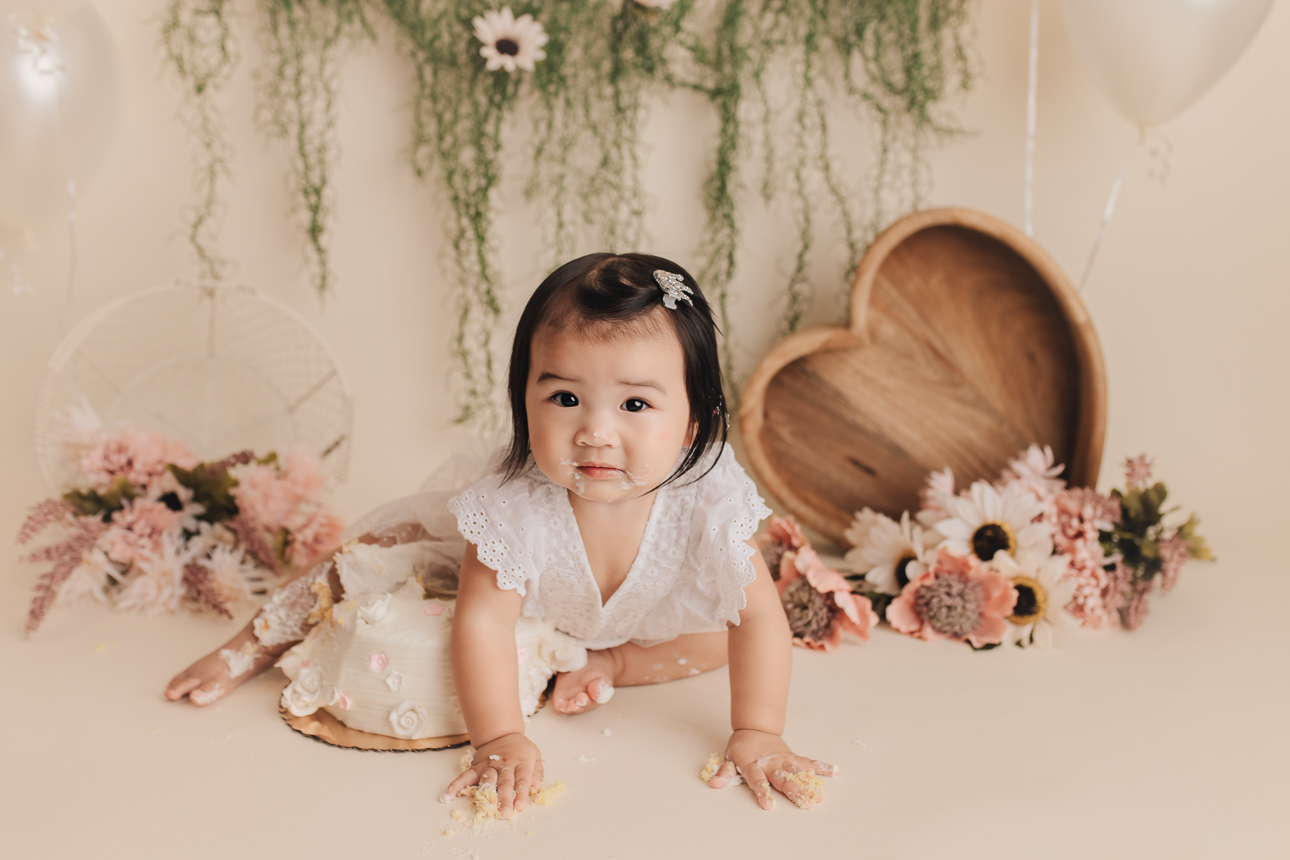 Baby girl with frosting on face crawling over cake during a pink-and-white floral cake smash session with beige backdrop.