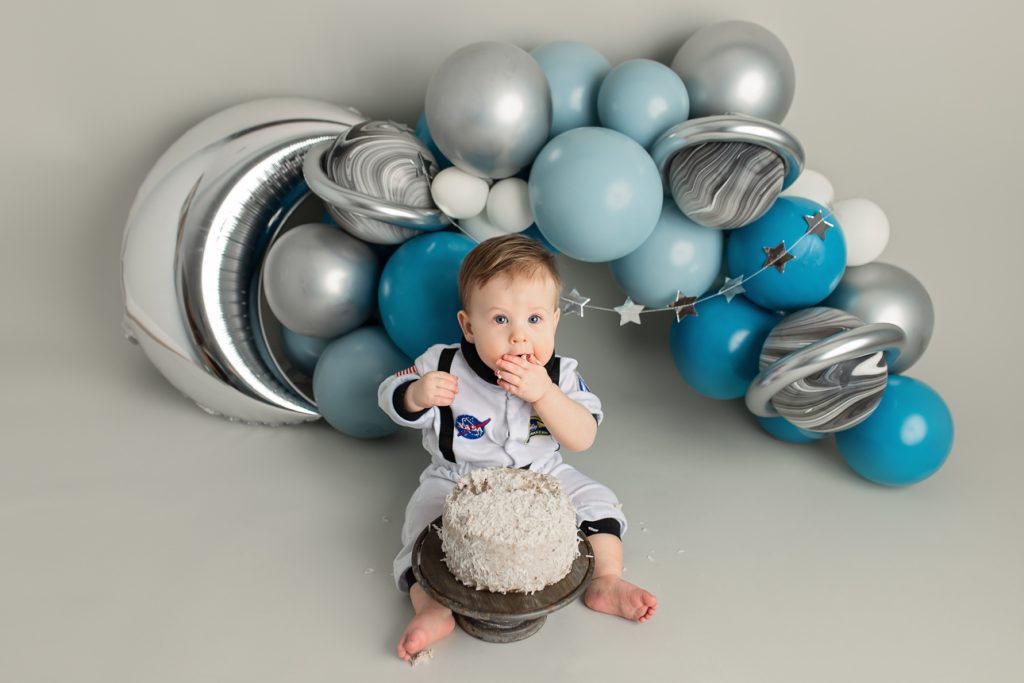 Baby boy in astronaut outfit eating cake during outer space themed cake smash with balloon arch, moon, stars and planets.