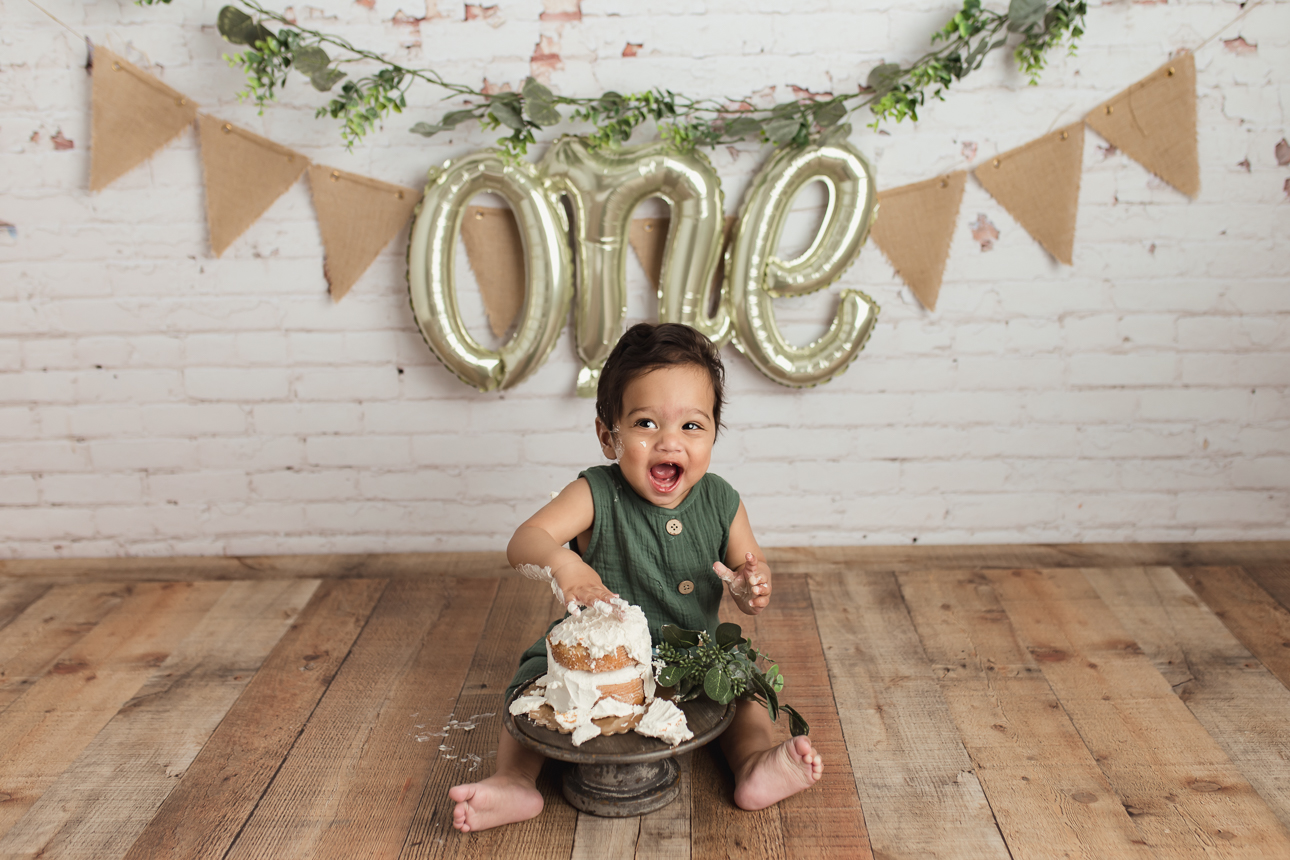 Baby boy in green romper laughing and digging into cake during boho cake smash with greenery and rustic brick wall backdrop.