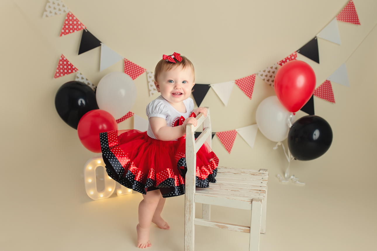 Baby girl in red and black tutu dress standing holding onto a small chair during cake smash; garland and balloons behind.