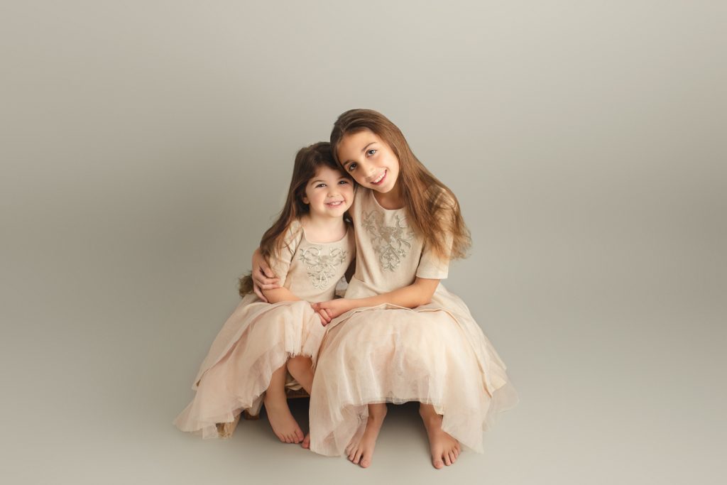 School-aged sisters in matching pink dresses, sitting barefoot and smiling while holding hands on a gray studio backdrop.