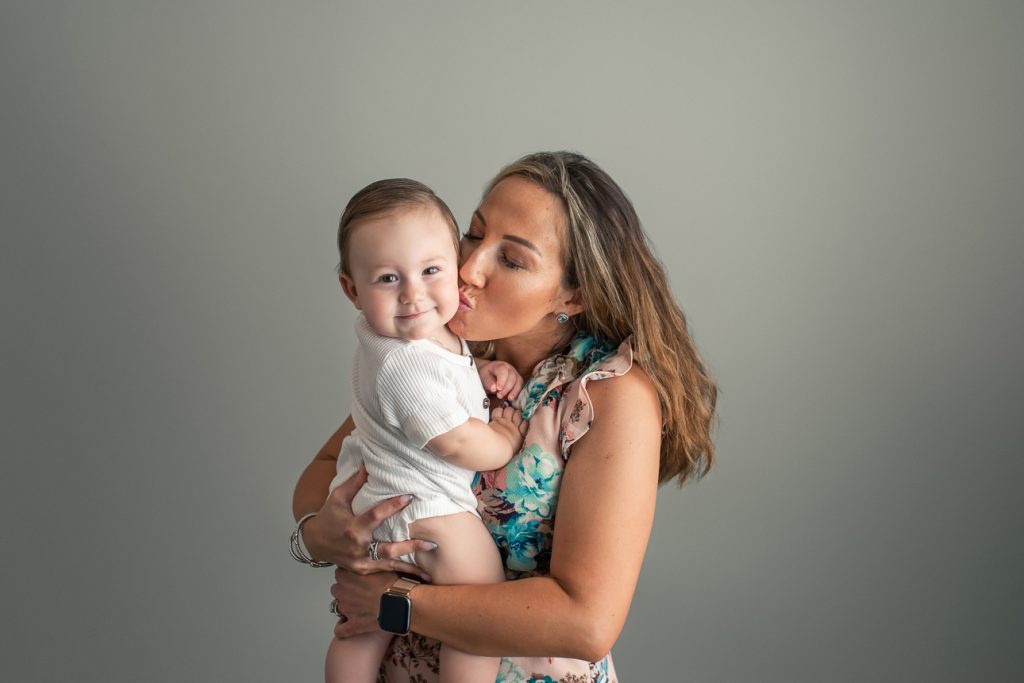 Mom squeezing and kissing baby boy on cheek while he looks and smiles at the camera during a studio milestone photo session.