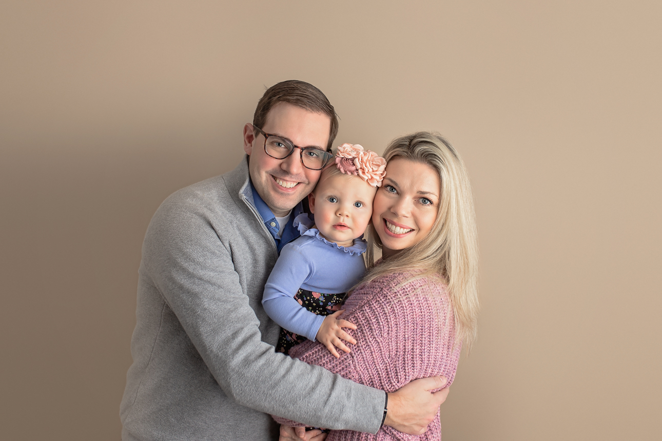Studio milestone portrait of parents embracing baby girl in center, all smiling at camera; on a light brown backdrop.