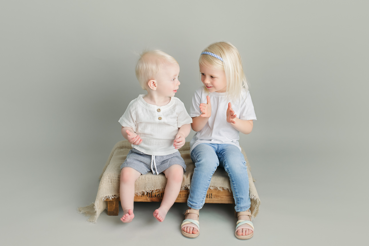Toddler brother and preschool sister dressed in white sit on bench clapping and smiling at each other in a studio portrait.