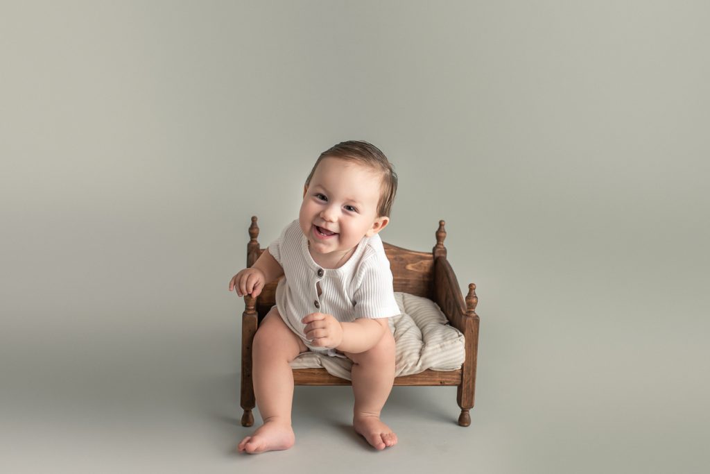 Laughing baby boy in white romper sitting on little wooden bed prop with grey backdrop during a studio milestone photoshoot.