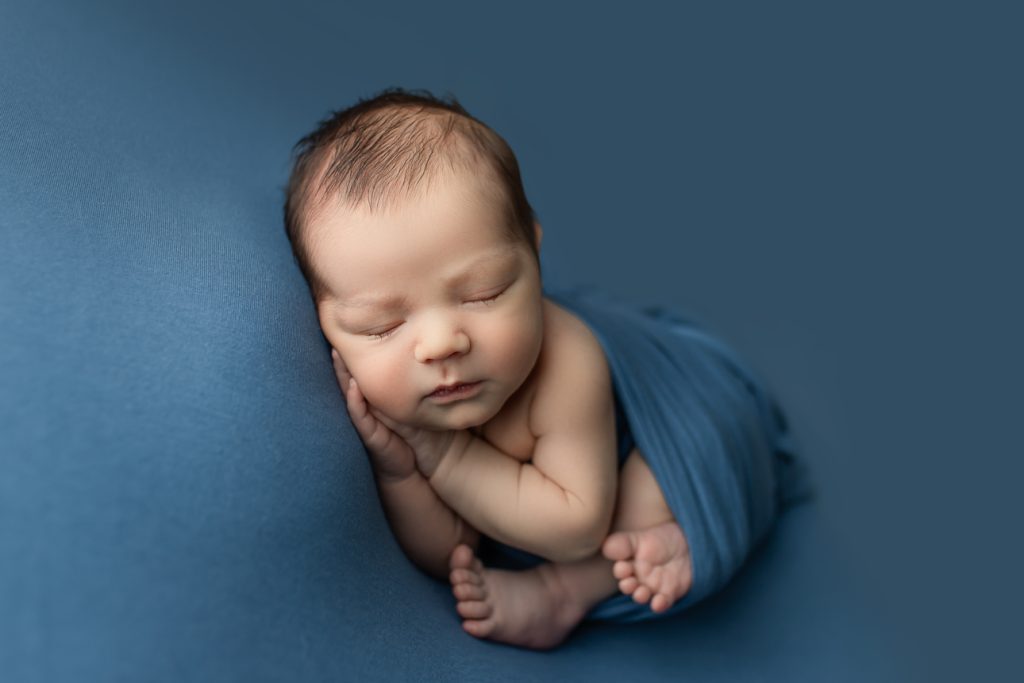 Newborn on blue backdrop, wrapped with arms and legs visible, sleeps on side with hands under cheek; studio session.