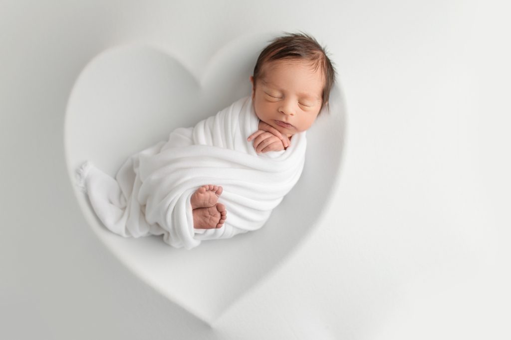 Newborn baby wrapped criss-cross style with feet showing on white backdrop, heart-shaped outline around baby; studio session.