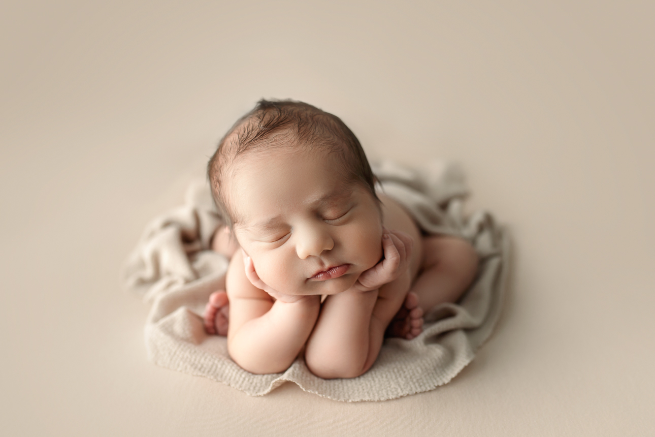 Newborn baby in froggy pose, arms propped under head and feet sticking out, resting on a soft beige backdrop; studio session.