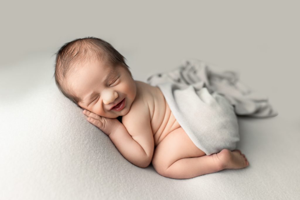 Newborn boy in bum-up pose sleeping on soft grey backdrop with a slight grin and skin rolls on back; studio newborn session.
