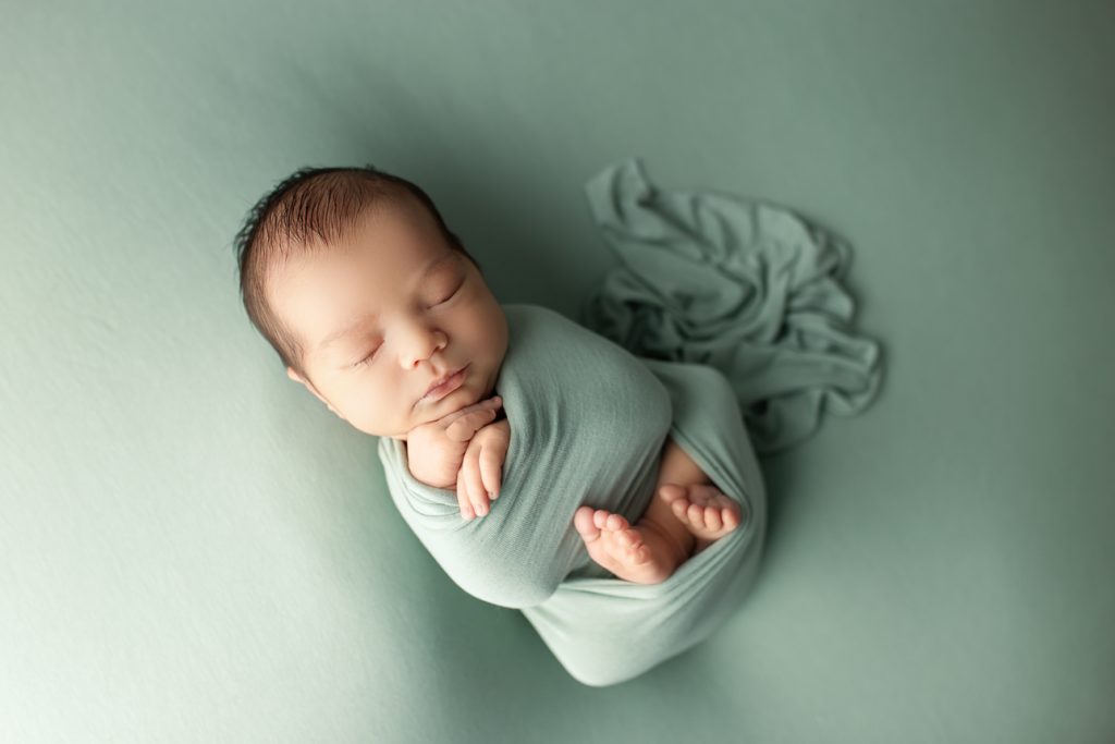 Sleeping newborn wrapped in pastel green, posed with hands under chin and tiny feet peeking out; peaceful studio portrait.