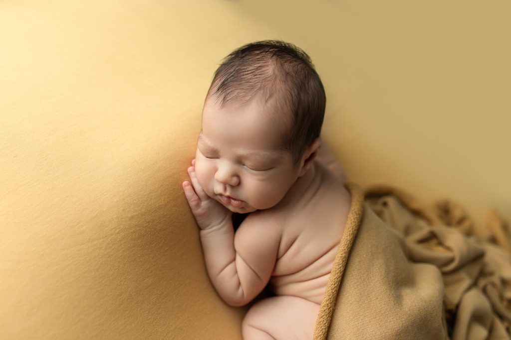 Sleeping newborn baby posed tummy down on a soft yellow backdrop, hand under cheek, showing adorable rolls and wrinkles.