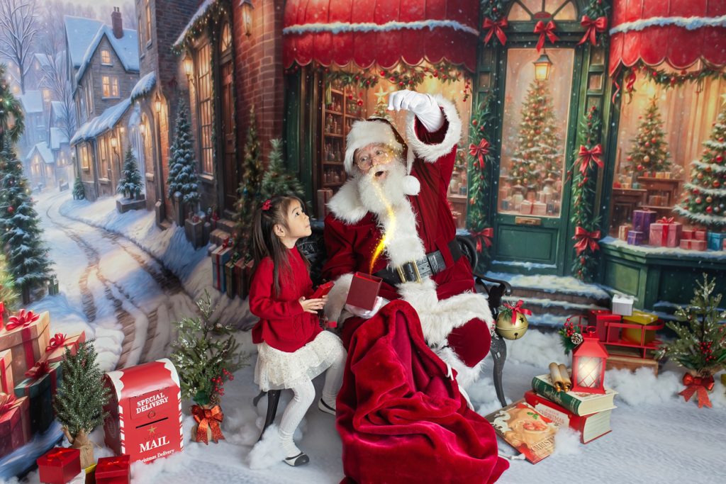 Santa sits with a young girl as she holds open a red gift box, with magic light shooting out, and she looks up in wonder.
