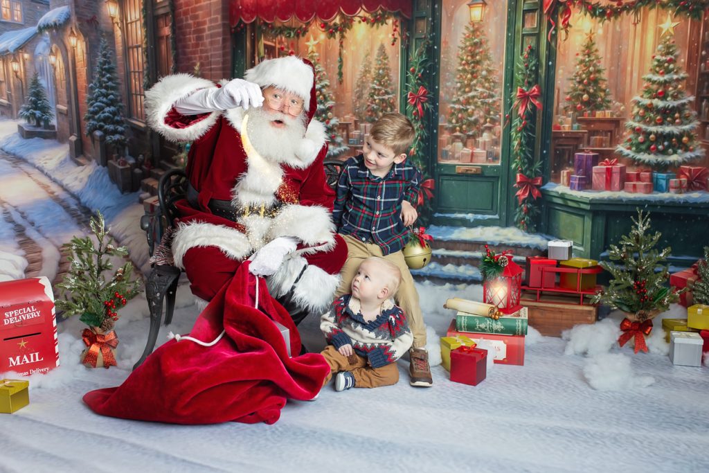 Two young brothers sit in snowy holiday studio scene; they look up in awe as Santa sprinkles magic dust over his red toy bag.