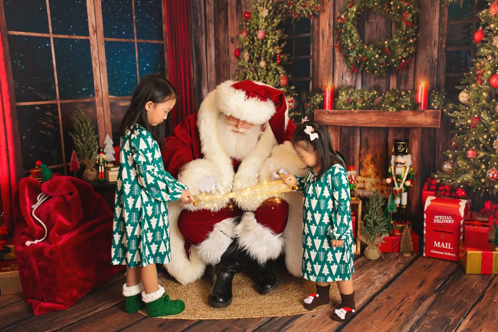Rustic holiday studio scene: two young girls in matching green pajamas stand with Santa looking at a glowing scroll.