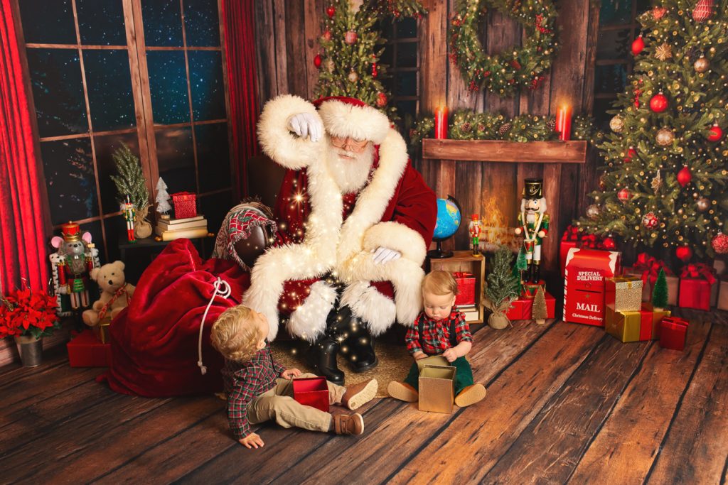 Two young brothers sit with Santa in holiday studio scene; they watch in awe as Santa sprinkles magic dust over gift boxes.