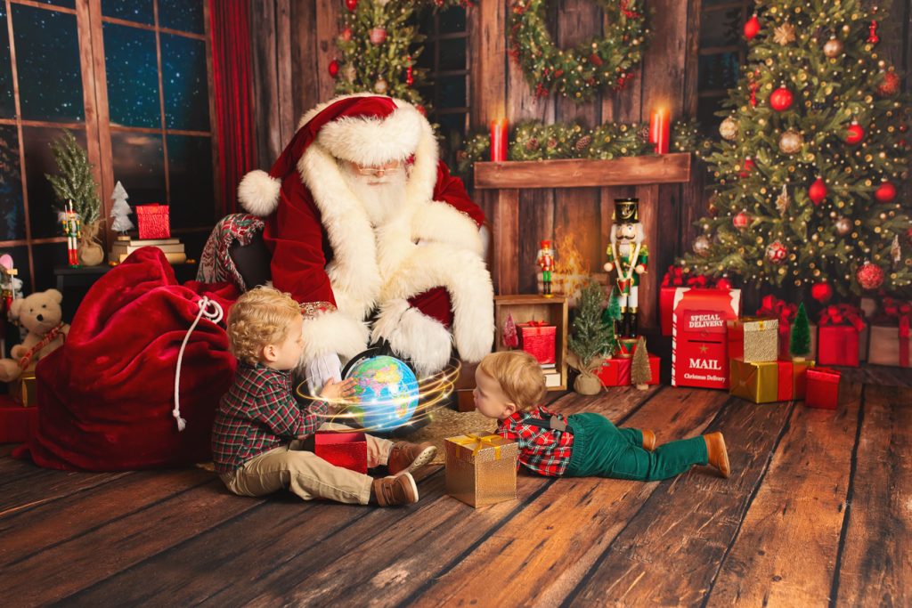 Rustic holiday studio scene: two young brothers sitting with Santa, one spins a glowing globe while the other watches in awe.