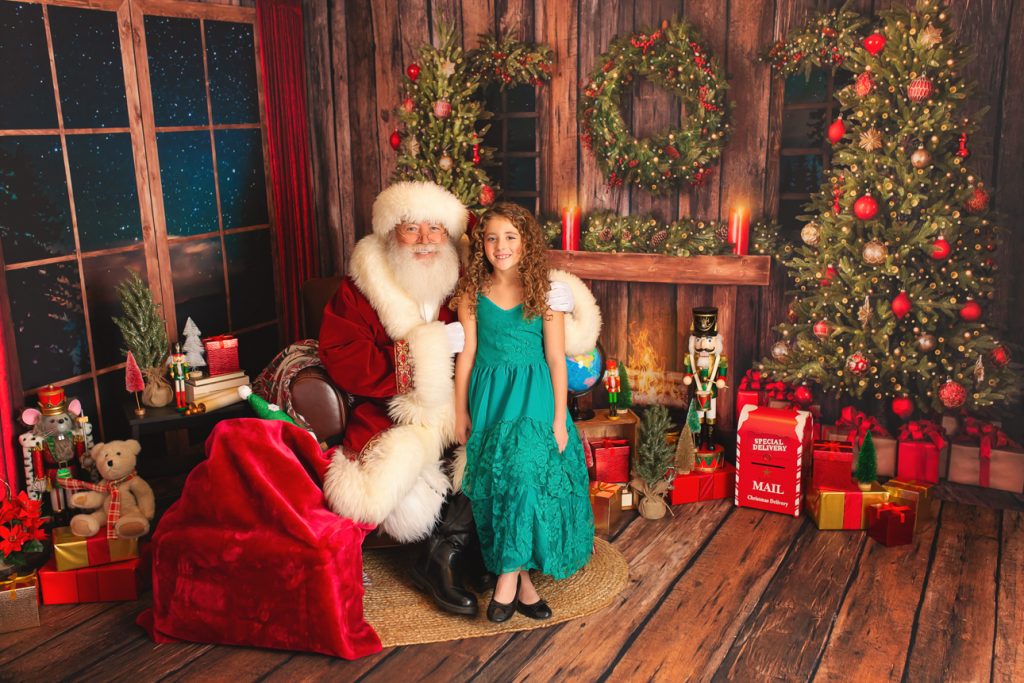 Rustic indoor holiday studio scene; Santa sits with a young girl in a green dress, both smiling warmly at the camera.