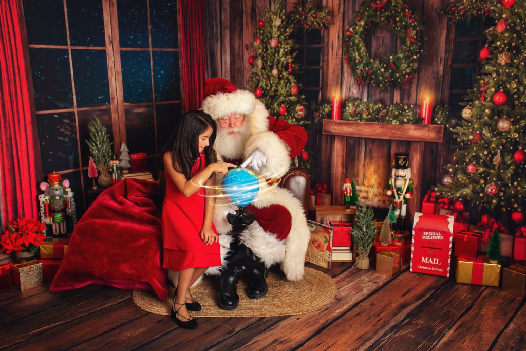 Rustic holiday studio scene: young girl in a red dress sits with Santa, both pointing at a magic spinning globe.